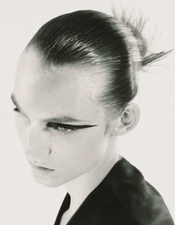 Close-up portrait of a model with sleek pulled-back hair and dramatic winged eyeliner against a plain background.