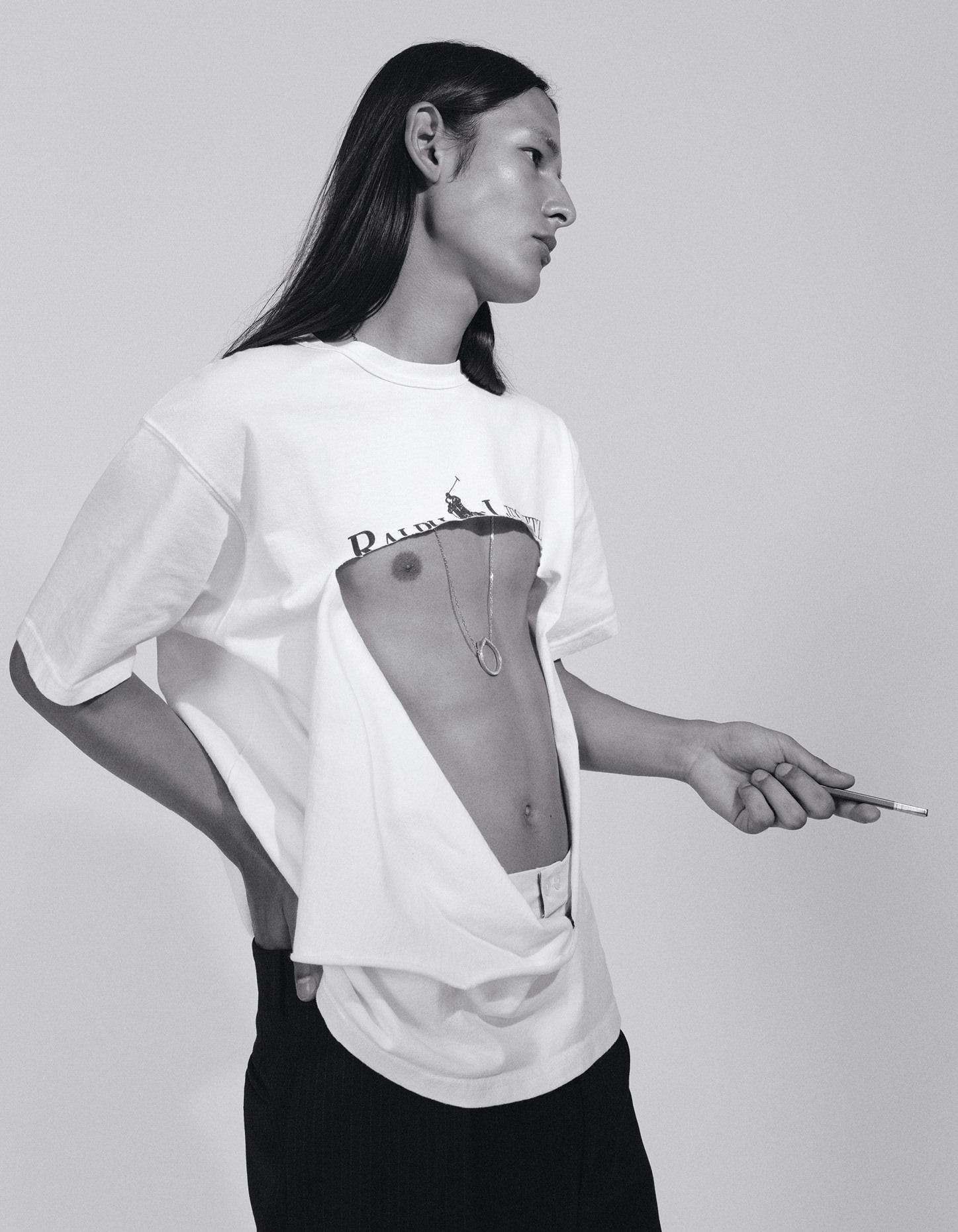 Monochrome portrait of androgynous person in deconstructed shirt with cigarette, striking a pose against a stark white backdrop.