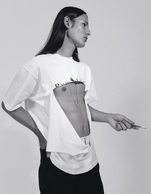 Monochrome portrait of androgynous person in deconstructed shirt with cigarette, striking a pose against a stark white backdrop.