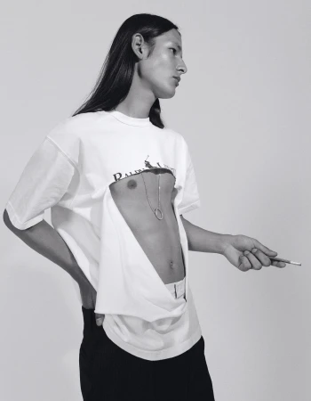 Monochrome portrait of androgynous person in deconstructed shirt with cigarette, striking a pose against a stark white backdrop.