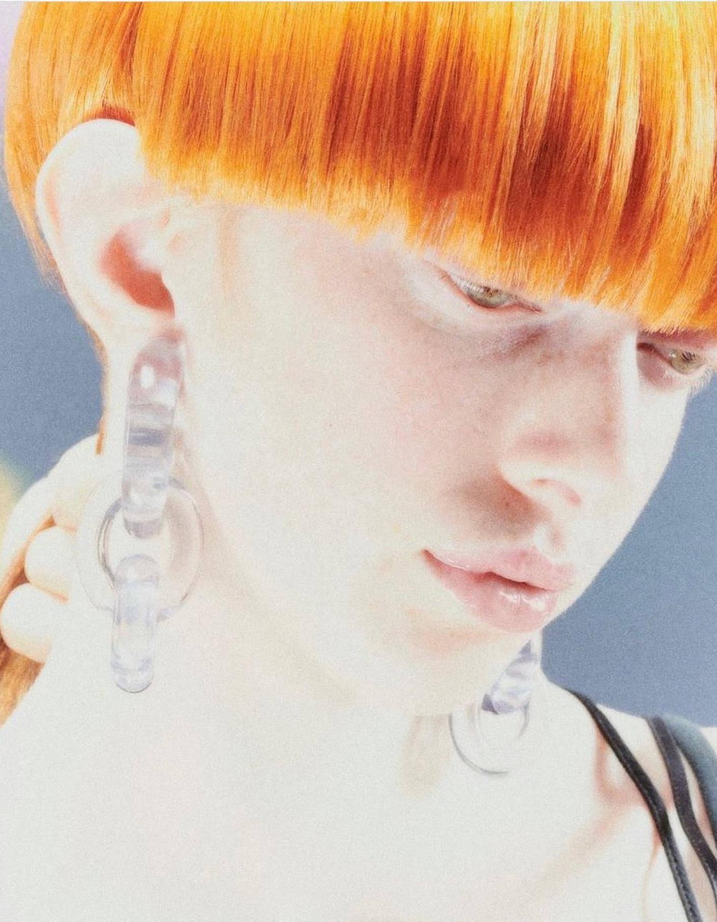 Close-up portrait of an adult female with orange hair wearing clear hoop earrings, against a blurred blue and yellow background.
