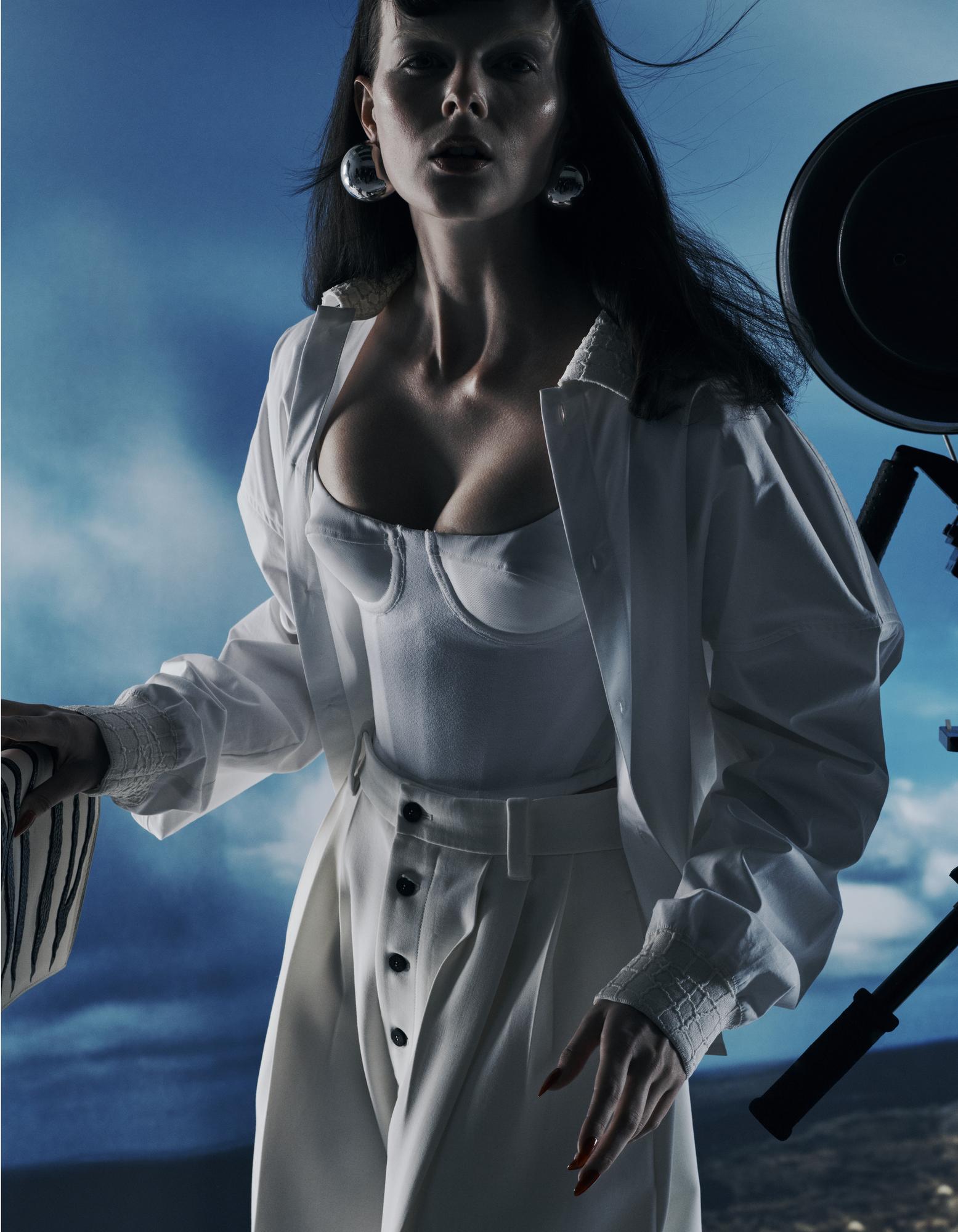 A woman in a white top and pants poses outdoors with camera equipment. The model has dark hair and is wearing statement earrings.