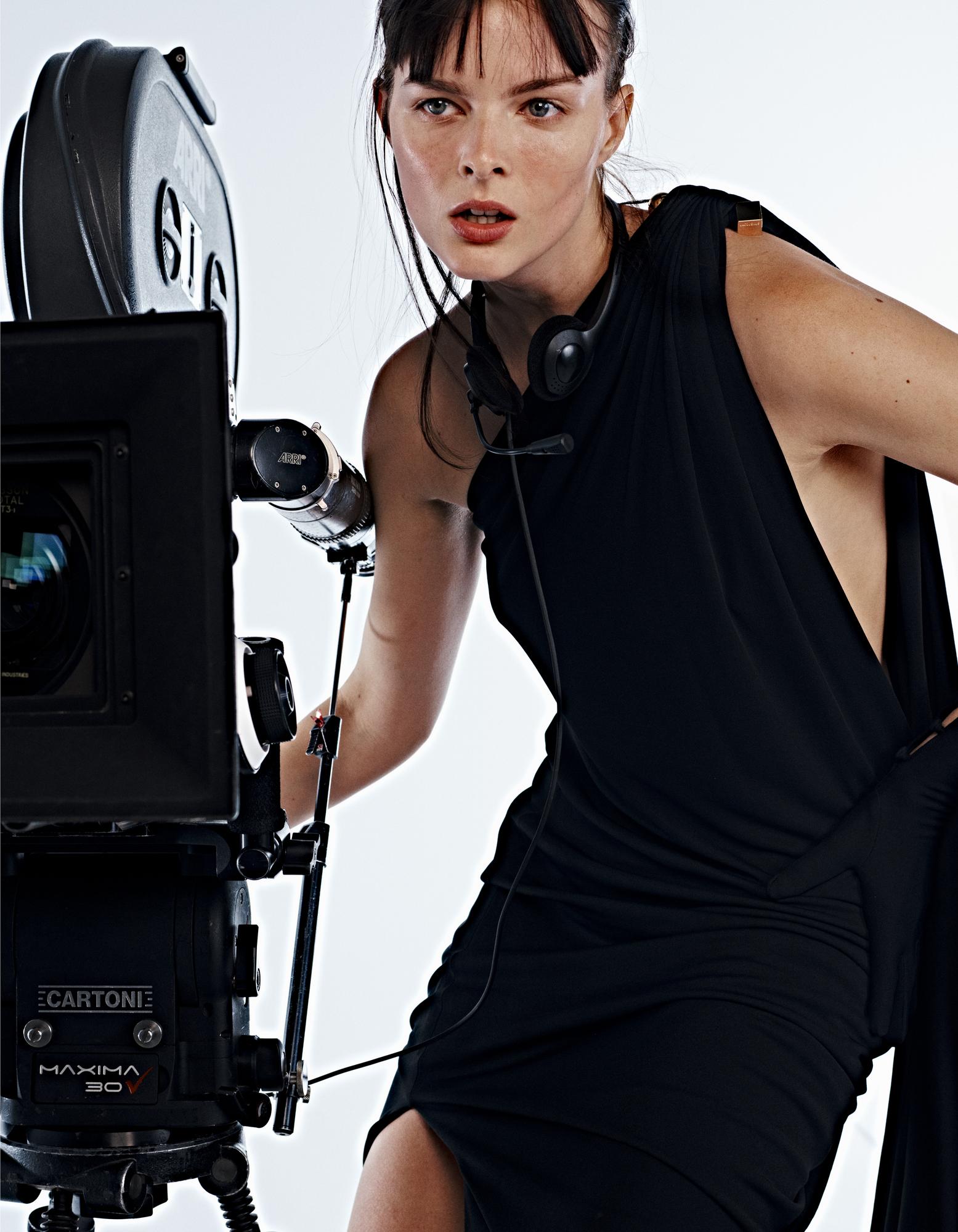 Woman in black dress poses with a vintage film camera in a studio setting, creating a striking fashion image.