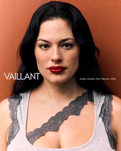 Portrait of Ashley Graham in a grey lace-trimmed tank top against a textured brown background, showcasing her dark hair and red lipstick.