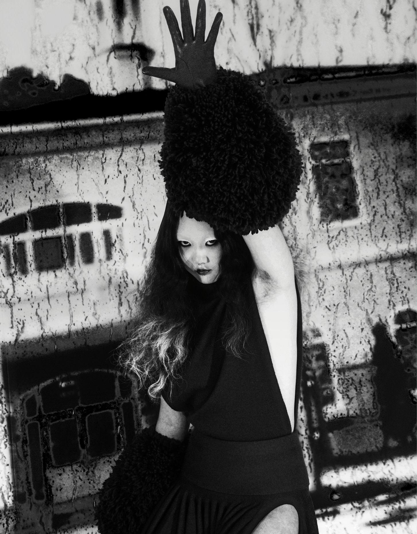 Black and white fashion shot of a woman in black with textured arm coverings and hand raised, with a blurred urban wall background.