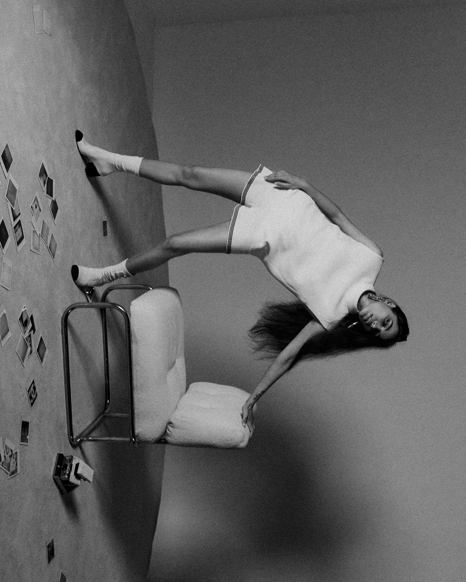 Artistic black-and-white image of a woman lying on a chair against a wall decorated with photos, showcasing creative composition.