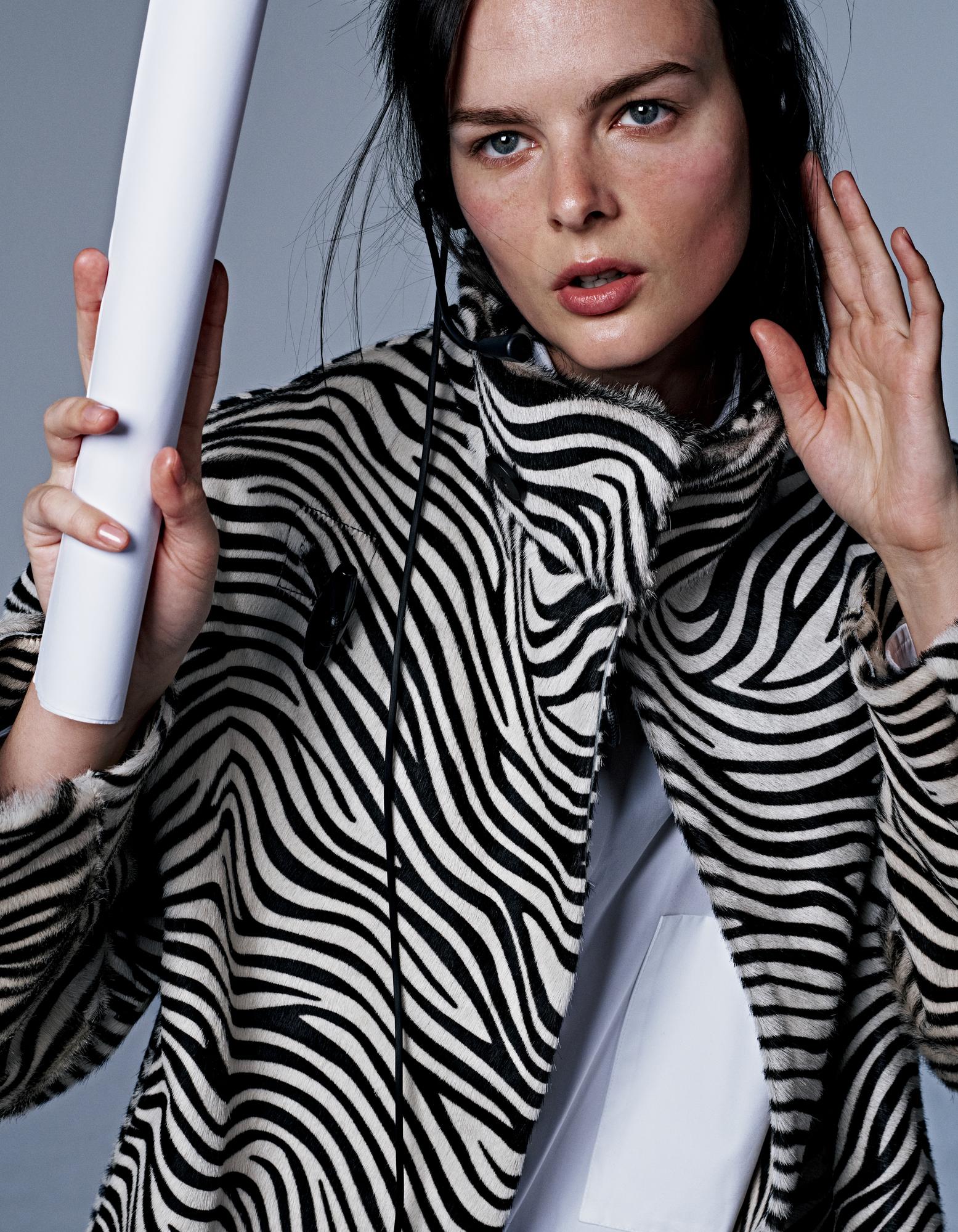Fashionable woman in a zebra print coat holds a white tube in a studio setting with a light gray background, emphasizing modern style.