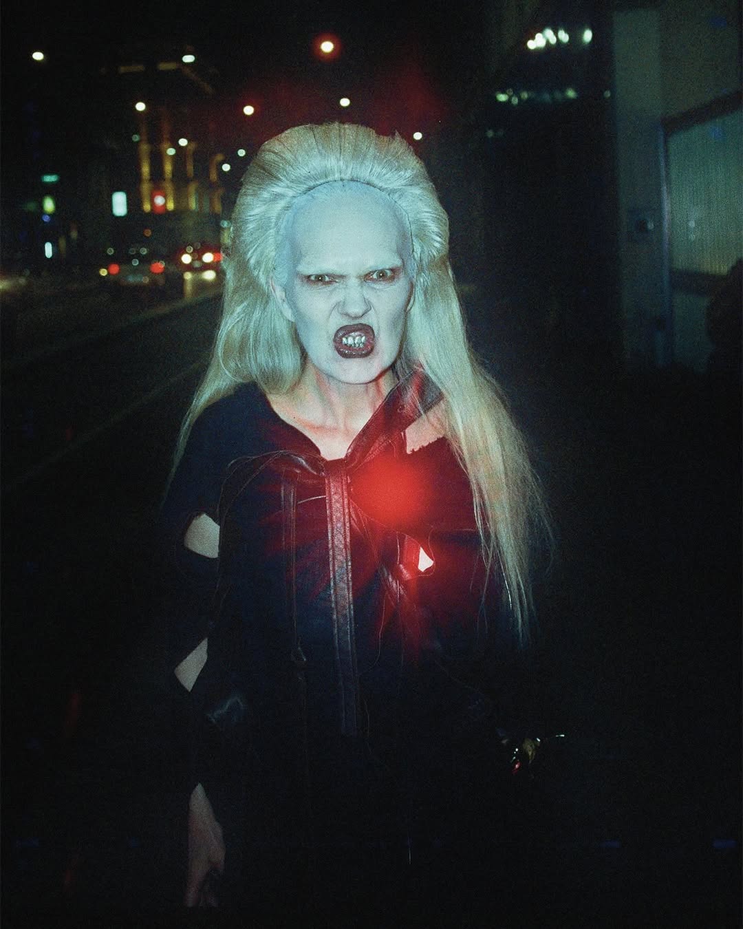 A person with pale makeup and white hair grimaces in a black jacket on a city street at night with vehicle lights visible in the background.
