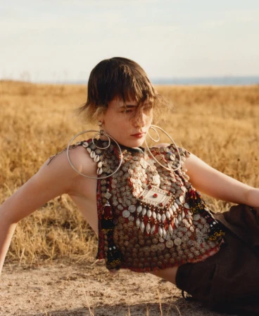 Model wearing ornate coin-embroidered top and large silver hoop earrings resting on dry grassy terrain under warm sunlight.
