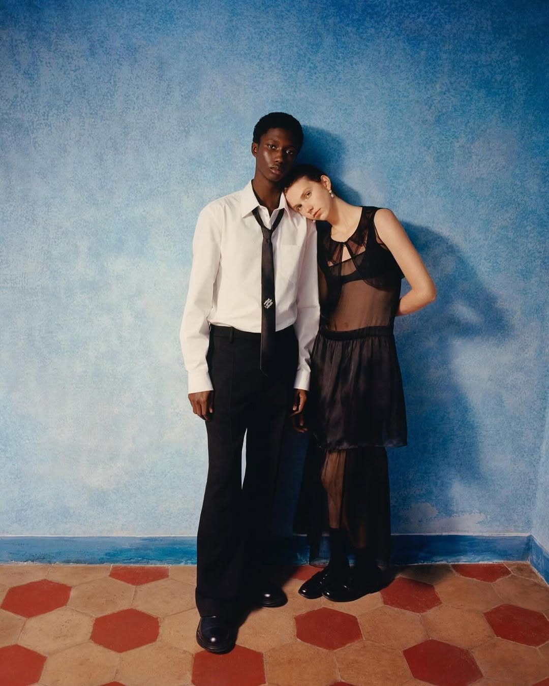 A slender man and woman pose together in a fashion shot, the woman resting her head on the man's shoulder against a textured blue wall.