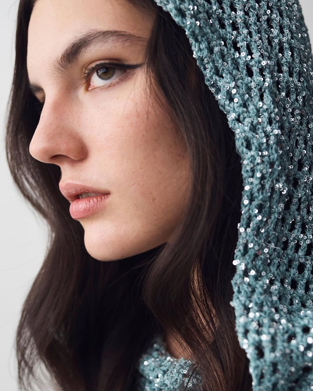 Close-up portrait of a young woman with dark hair, wearing a shimmering teal crochet hooded top, set against a neutral backdrop. Beauty and fashion.