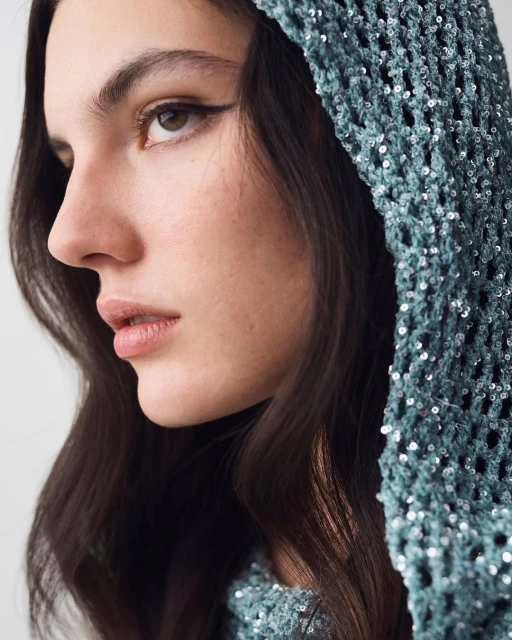 Close-up portrait of a young woman with dark hair, wearing a shimmering teal crochet hooded top, set against a neutral backdrop. Beauty and fashion.