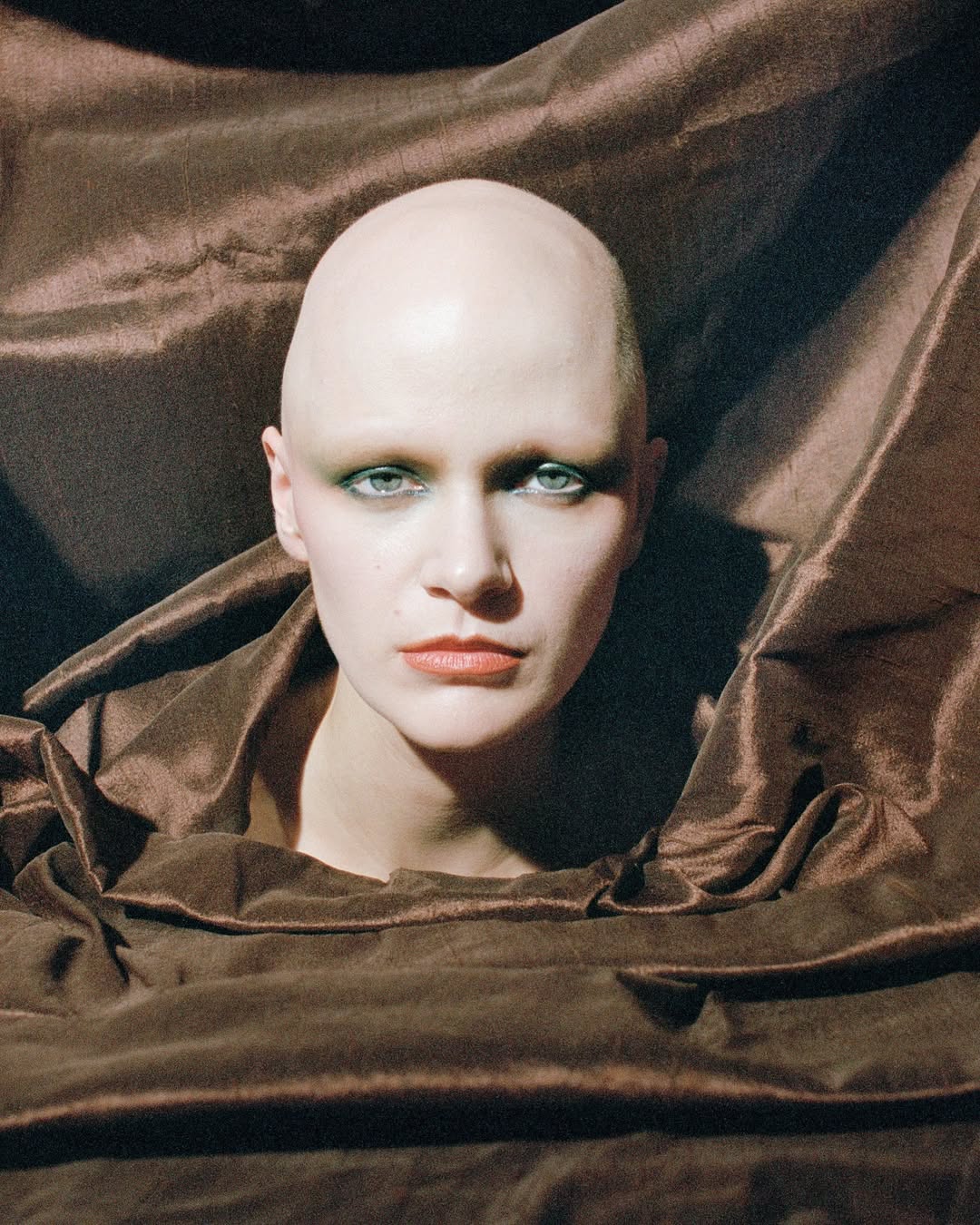 A portrait of a bald adult woman with green eye makeup, wearing a brown fabric collar, against a brown draped fabric backdrop.