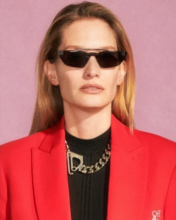 Close-up portrait of a woman wearing black angular sunglasses, a vibrant red blazer, and a chunky silver chain necklace.
