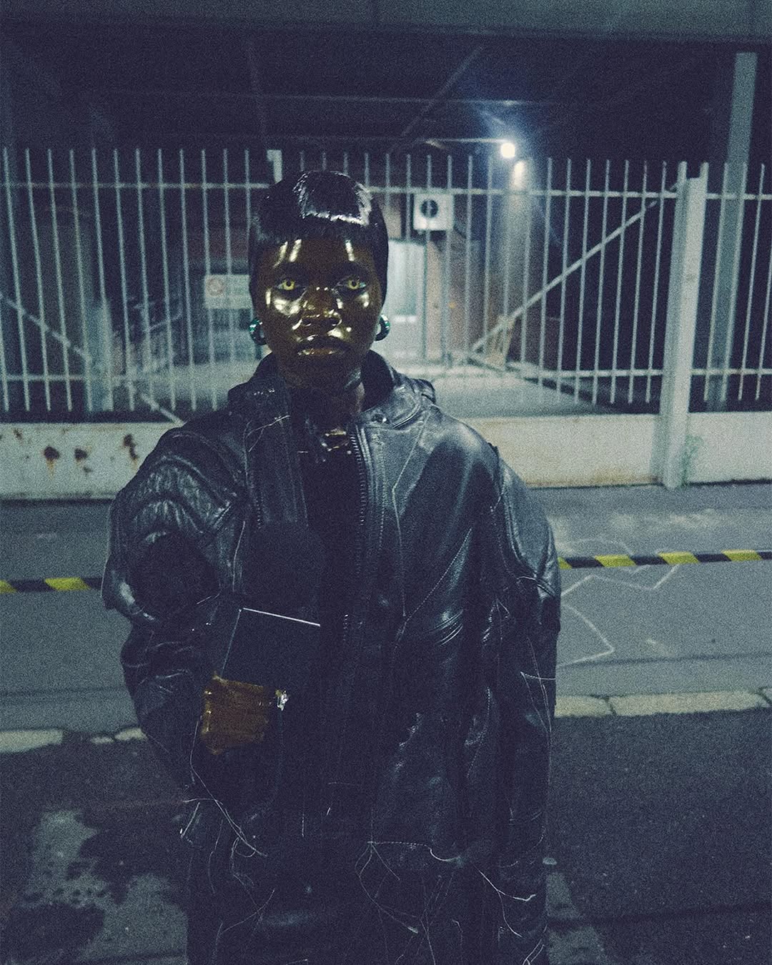 A person with dark skin wearing a black jacket and green earrings stands in front of a metal fence, illuminated by dim lighting.