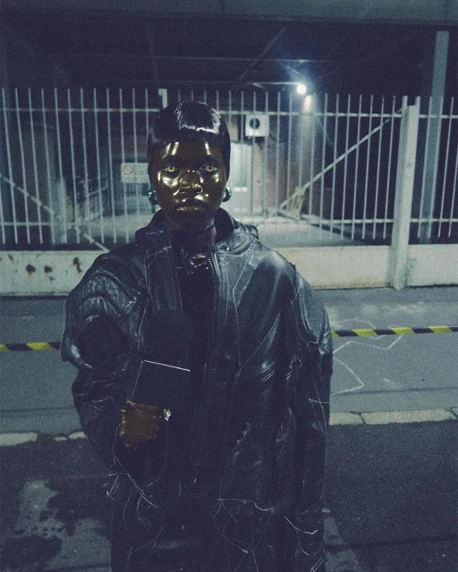 A person with dark skin wearing a black jacket and green earrings stands in front of a metal fence, illuminated by dim lighting.