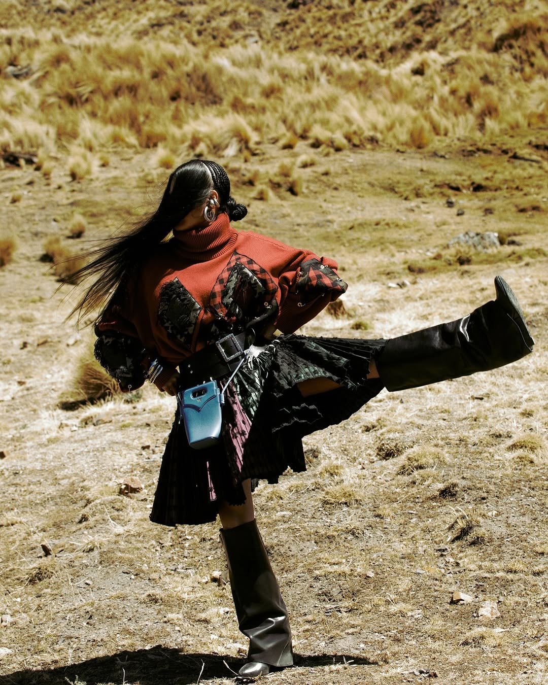 Woman in high boots and red patterned top poses outdoors in a kicking position near dried grass, showcasing fashion and artistic expression.