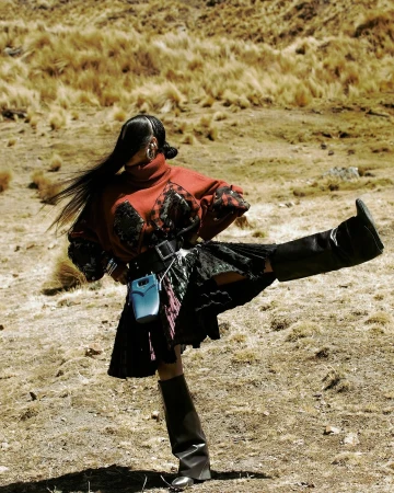 Woman in high boots and red patterned top poses outdoors in a kicking position near dried grass, showcasing fashion and artistic expression.