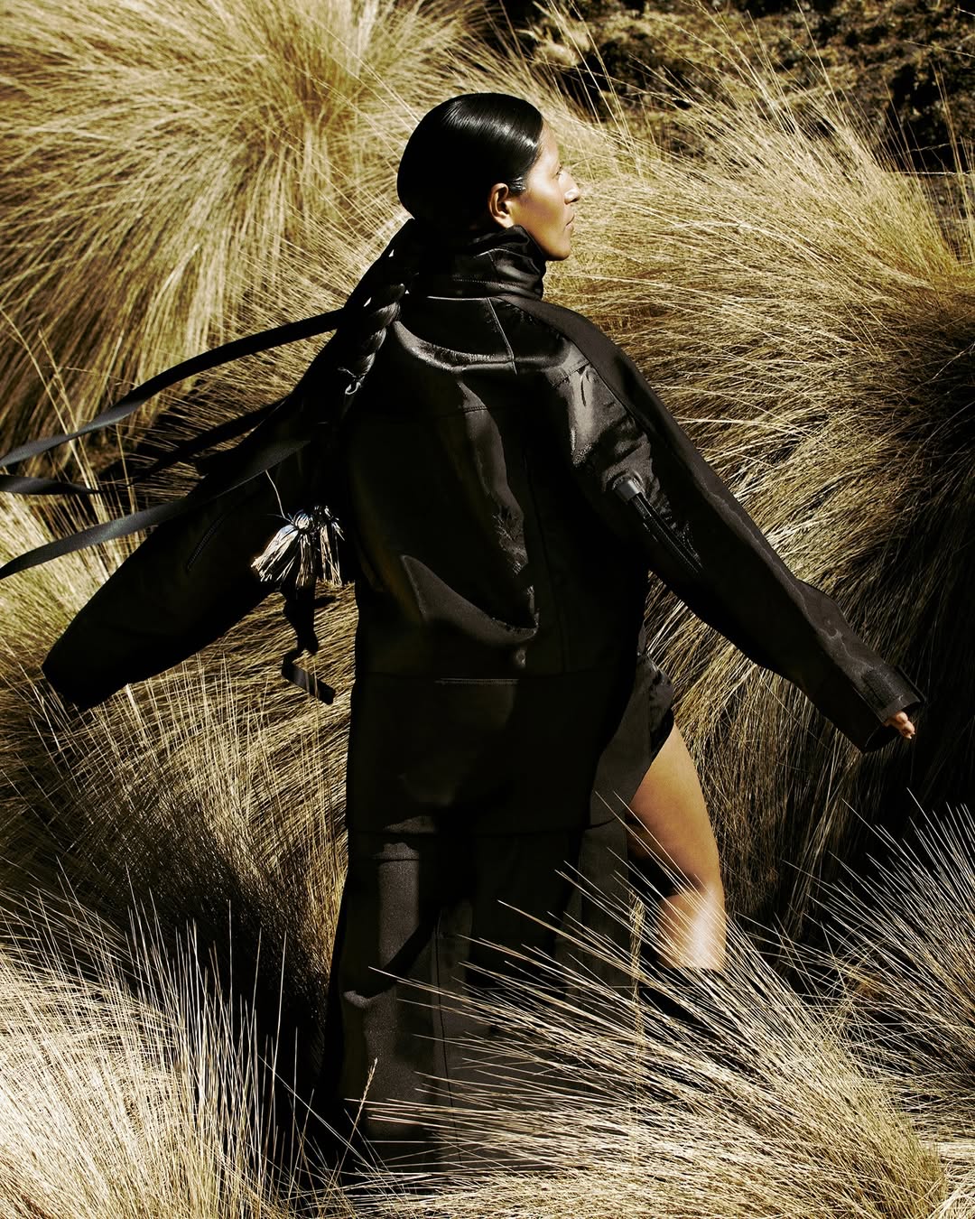 An adult female model stands in a field of tall grass wearing a dark jacket. The photograph is lit with natural sunlight.