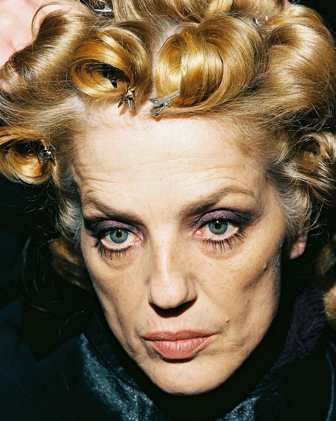 Close-up portrait of an adult woman with light blonde hair styled in curlers with metal clips, wearing dark eye makeup and a dark top.