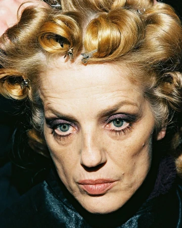 Close-up portrait of an adult woman with light blonde hair styled in curlers with metal clips, wearing dark eye makeup and a dark top.