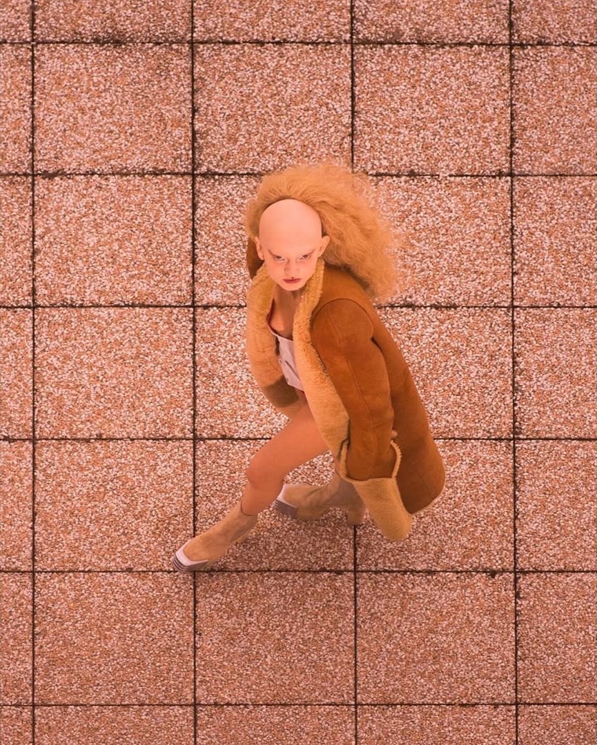 High-angle shot of a bald adult with blonde wig wearing a brown jacket and boots, standing on pink tiled ground, fashion-forward and unique.