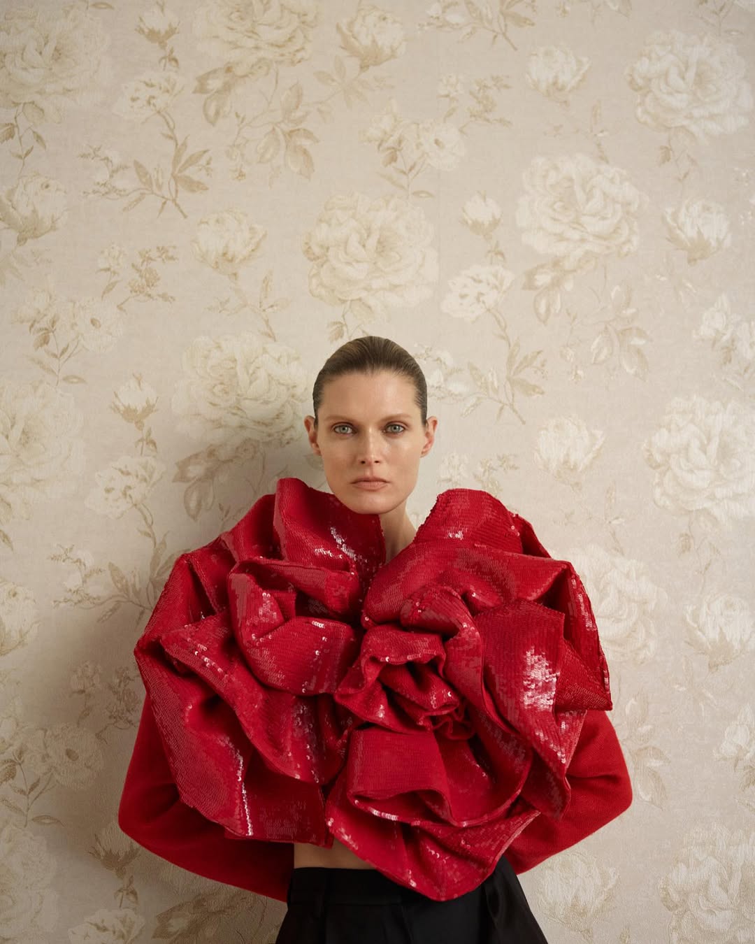 Fashion shot of an adult female wearing a red, sequined top with floral wallpaper in the background. Studio shot.