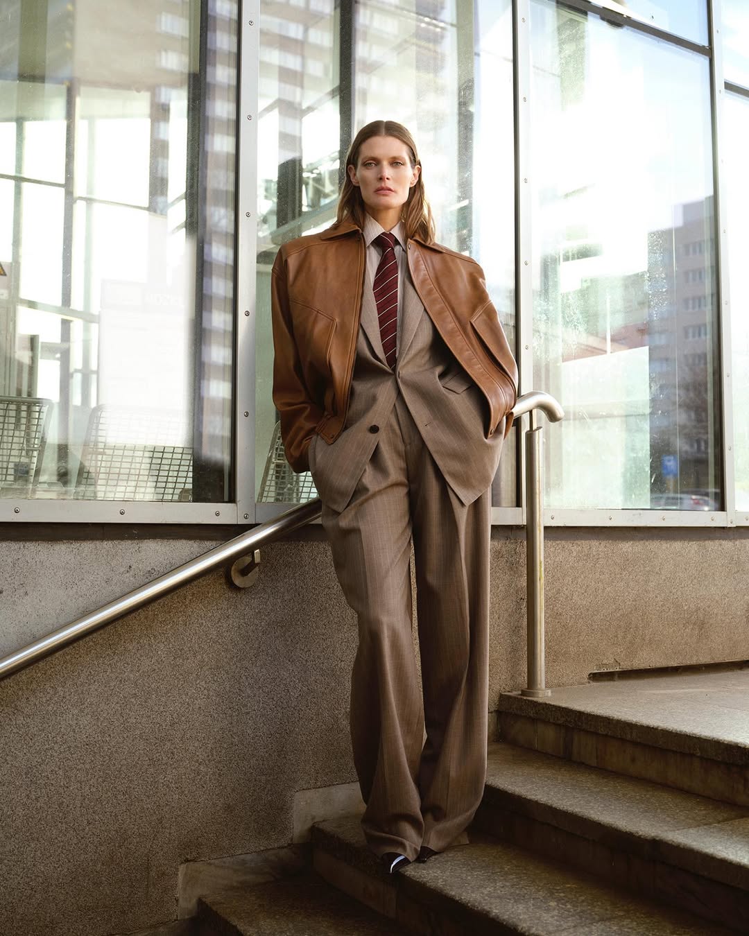 An adult female standing on concrete steps wearing a beige suit, brown leather jacket and striped tie in an urban setting.