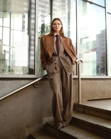 Model standing on stone steps leaning against a railing wearing a brown leather jacket and grey suit pants