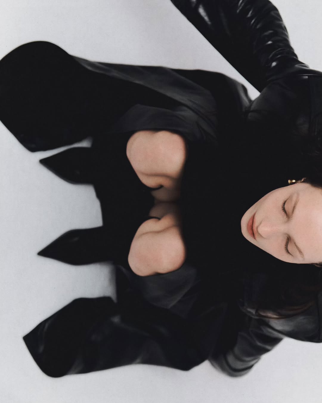 High angle fashion shot of a young woman with black coat and high boots posing in a white studio with soft, diffused lighting.