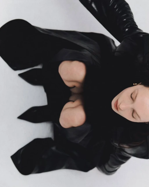 High angle fashion shot of a young woman with black coat and high boots posing in a white studio with soft, diffused lighting.