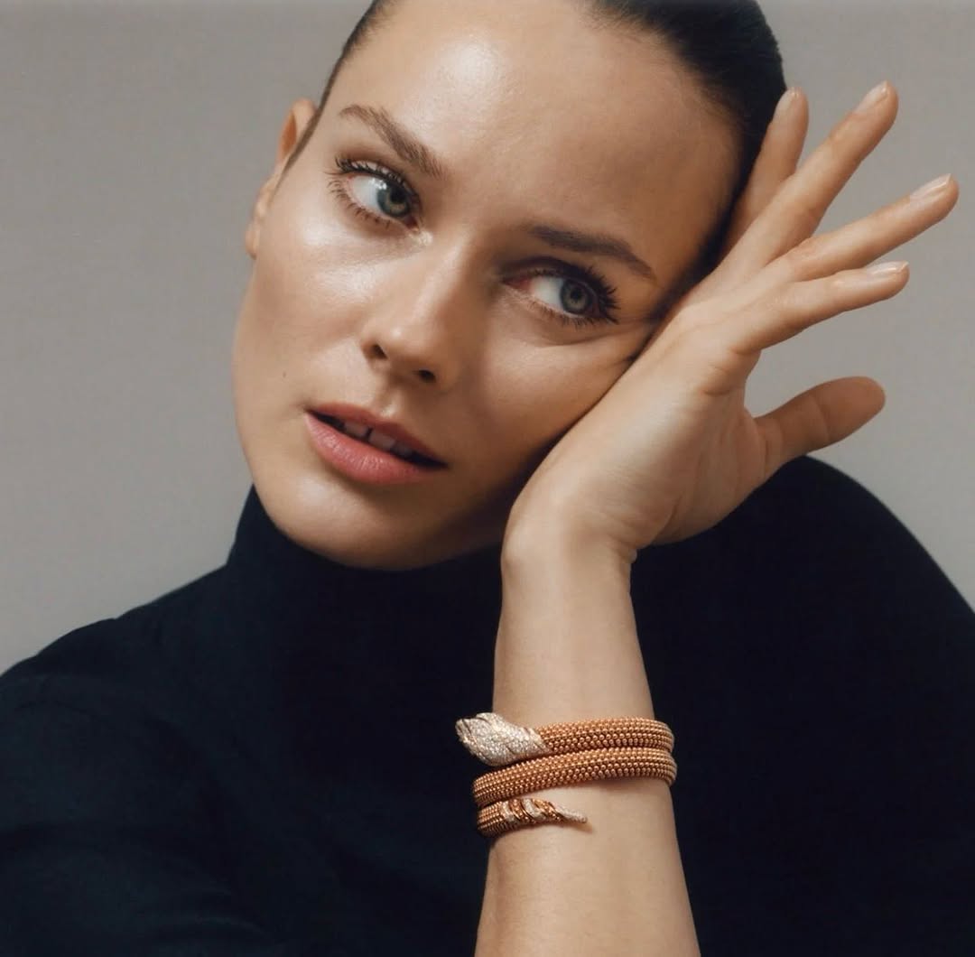 Close-up of a woman wearing a black turtleneck sweater and a golden snake bracelet, posing against a light gray backdrop in a studio.