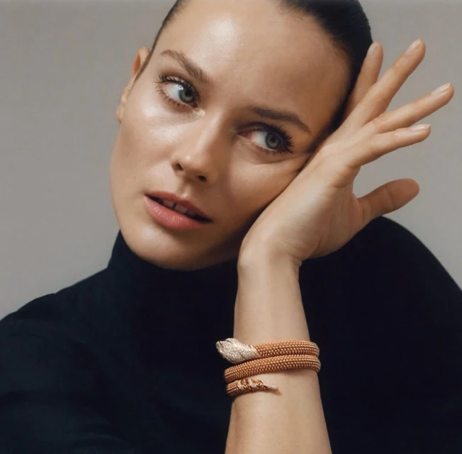 Close-up of a woman wearing a black turtleneck sweater and a golden snake bracelet, posing against a light gray backdrop in a studio.
