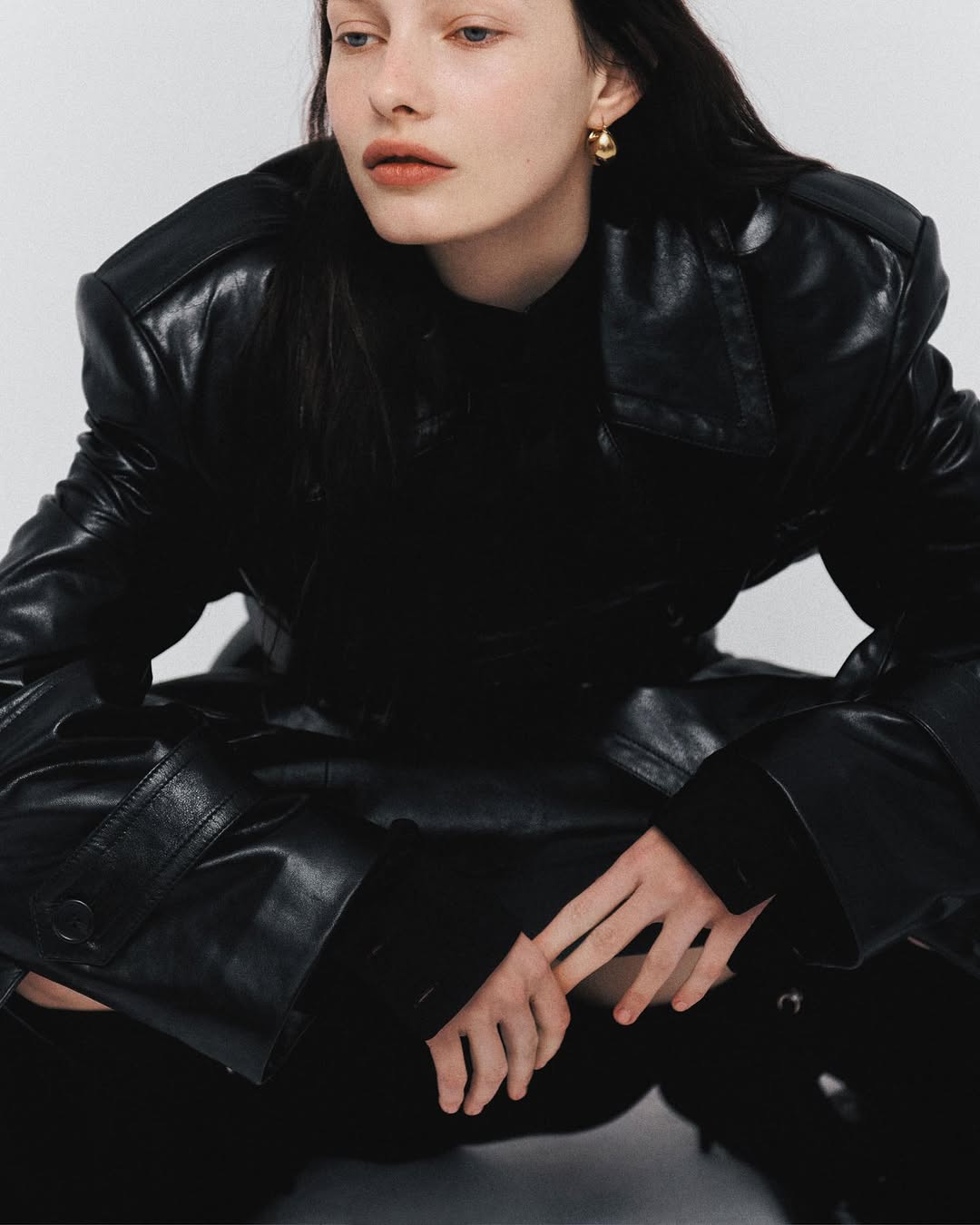 Elegant portrait of a young woman wearing a black leather jacket and gold earrings, captured in a photo studio against a white background.