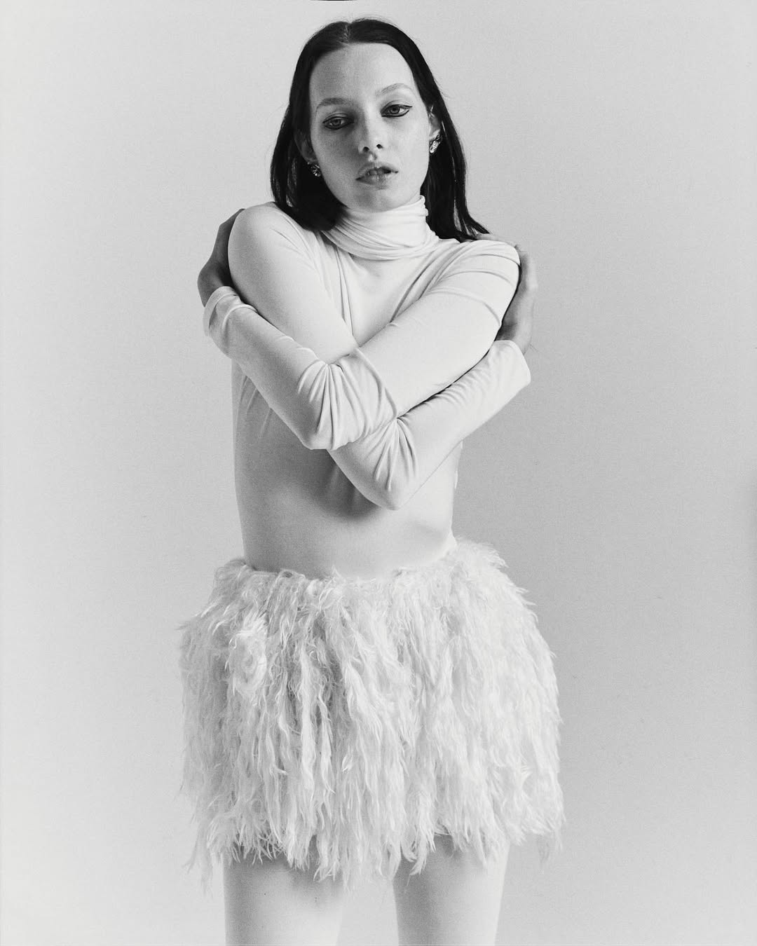 Monochrome portrait of a young adult woman with dark hair, wearing a white turtleneck and fluffy white miniskirt, posing indoors.