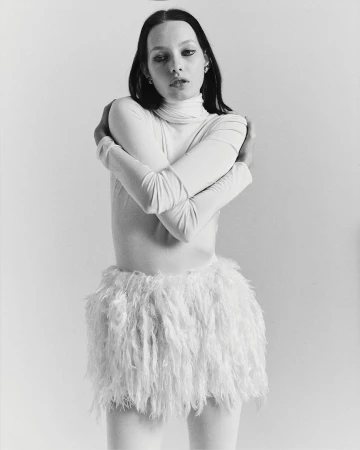 Monochrome portrait of a young adult woman with dark hair, wearing a white turtleneck and fluffy white miniskirt, posing indoors.