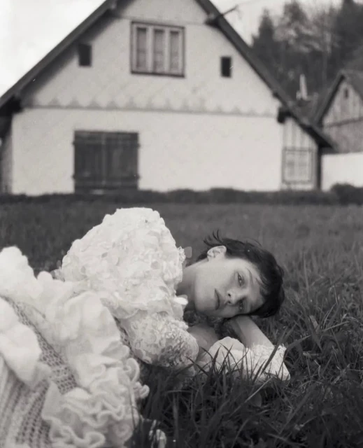 A young woman in a white dress lies in the grass in front of a white house in a black and white outdoor fashion portrait.