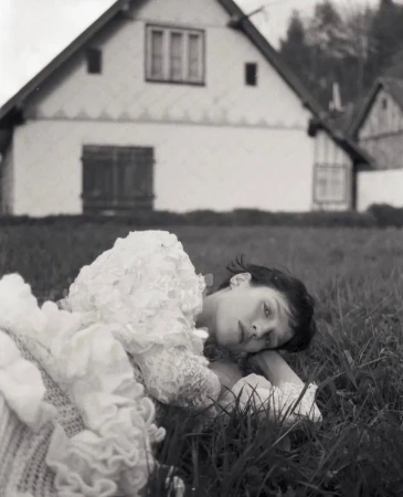 A young woman in a white dress lies in the grass in front of a white house in a black and white outdoor fashion portrait.