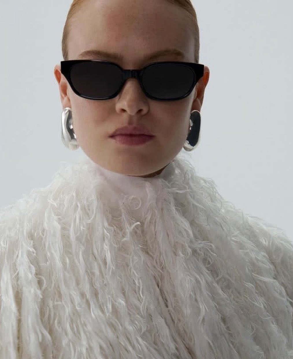 Close-up portrait of a woman wearing black sunglasses, silver earrings, and a textured white fringed top against a white backdrop.