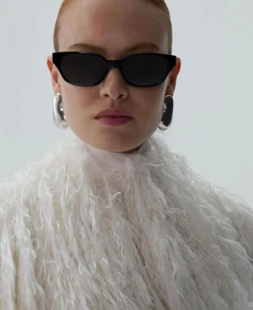 Close-up portrait of a woman wearing black sunglasses, silver earrings, and a textured white fringed top against a white backdrop.