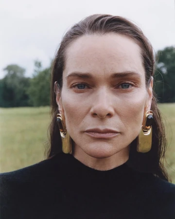 Close-up portrait of a woman with dark hair wearing statement gold earrings and a black top against a blurred outdoor background