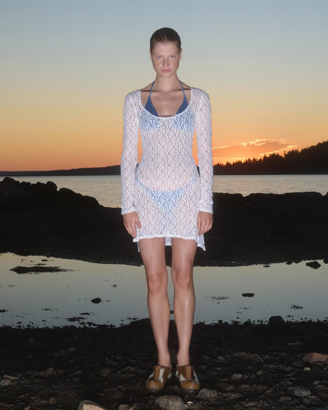 Full-shot of an adult female model in a white dress, standing on a rocky beach at sunset with the sky a gradient of orange and blue.