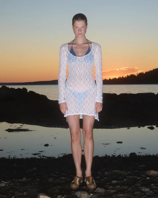 Full-shot of an adult female model in a white dress, standing on a rocky beach at sunset with the sky a gradient of orange and blue.