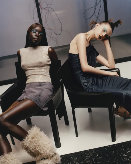 Two fashion models sit on black chairs indoors. A Black woman in a beige top and grey skirt sits next to a Caucasian woman in a blue dress.