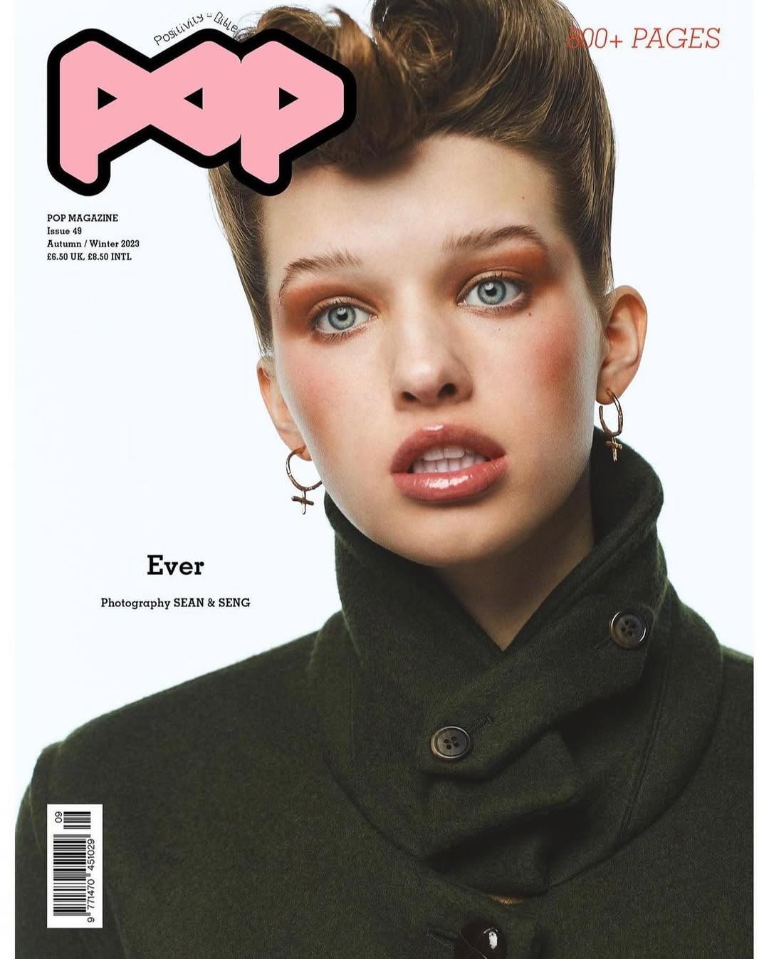 Fashion magazine cover featuring a woman with auburn hair and green coat, wearing hoop earrings with cross charms in a bright, studio setting.