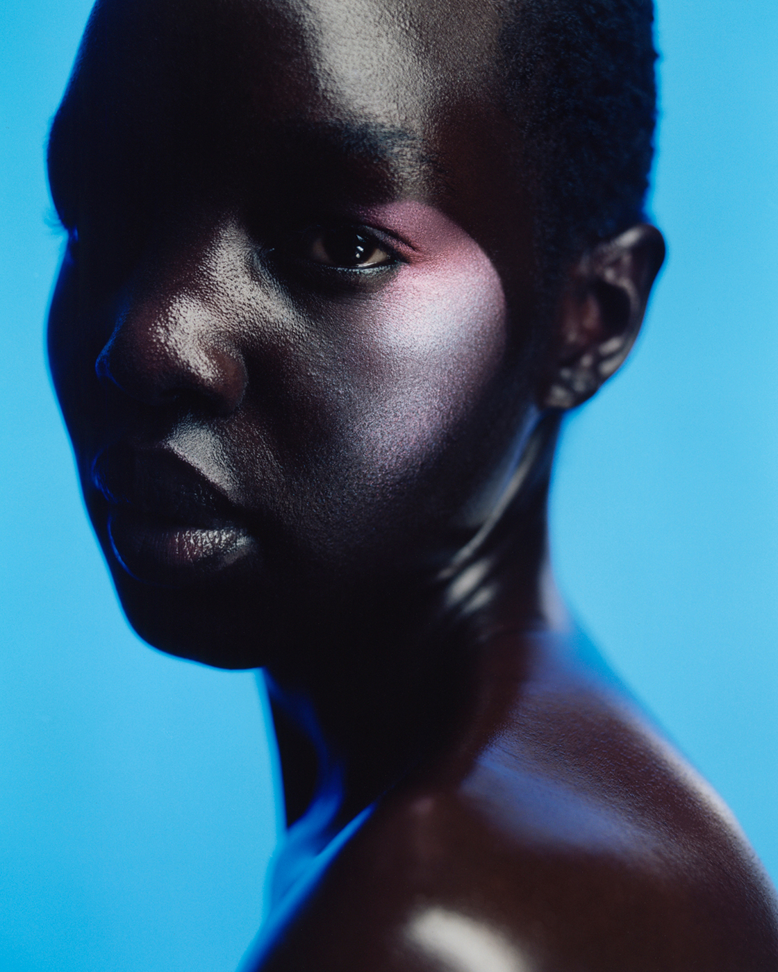 Close-up portrait of a dark-skinned adult with dramatic lighting and makeup against a vivid blue background in a studio setting.