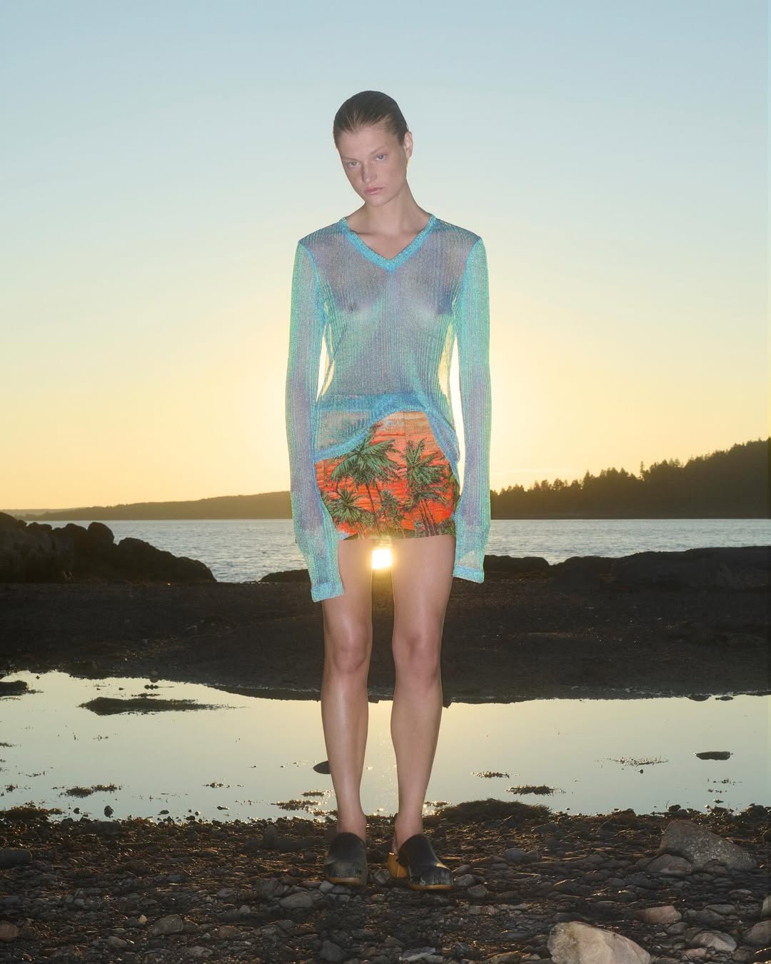 Model wearing a sheer long-sleeve blue top and tropical print skirt standing on a rocky shoreline at sunset by Julia Asaro