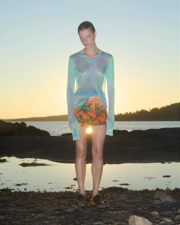 Model wearing a sheer long-sleeve blue top and tropical print skirt standing on a rocky shoreline at sunset by Julia Asaro