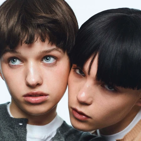 Close-up portrait of two young people with distinct dark hairstyles posing closely against a plain white background.