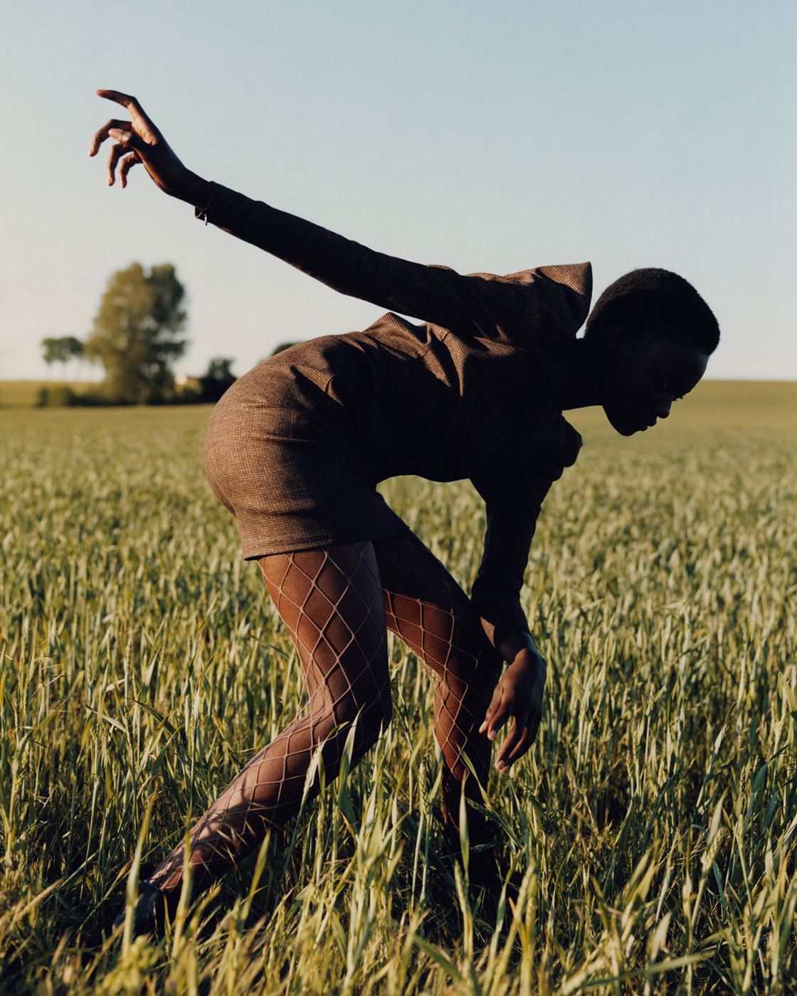 Woman in dress and fishnet stockings poses in a golden field, bending over with arms outstretched, under clear sky, fashion portrait.